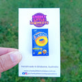 A hand holds a white display card featuring a miniature Cheezels box pin by Cheeky Little Monkeys Jewellery, with a blurred green grass background.