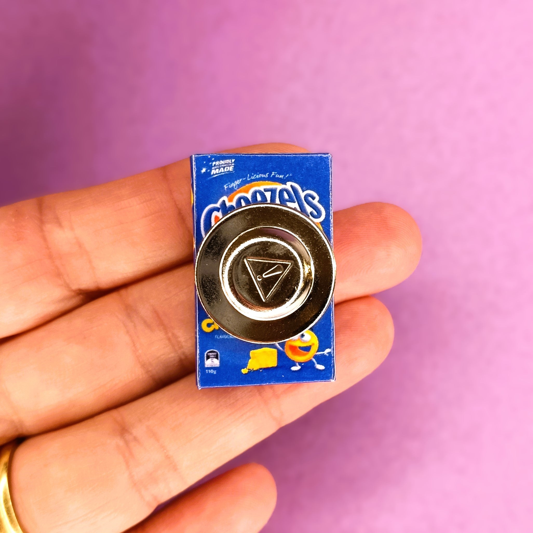 A small Cheetzels pin or brooch with a magnet back is held on a person's fingers against a solid pink background.