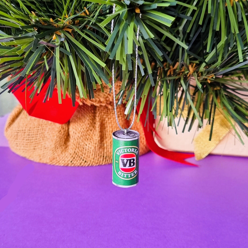 A miniature green and white VB Bitter beer can Christmas ornament hanging from a green artificial tree branch against a purple surface.