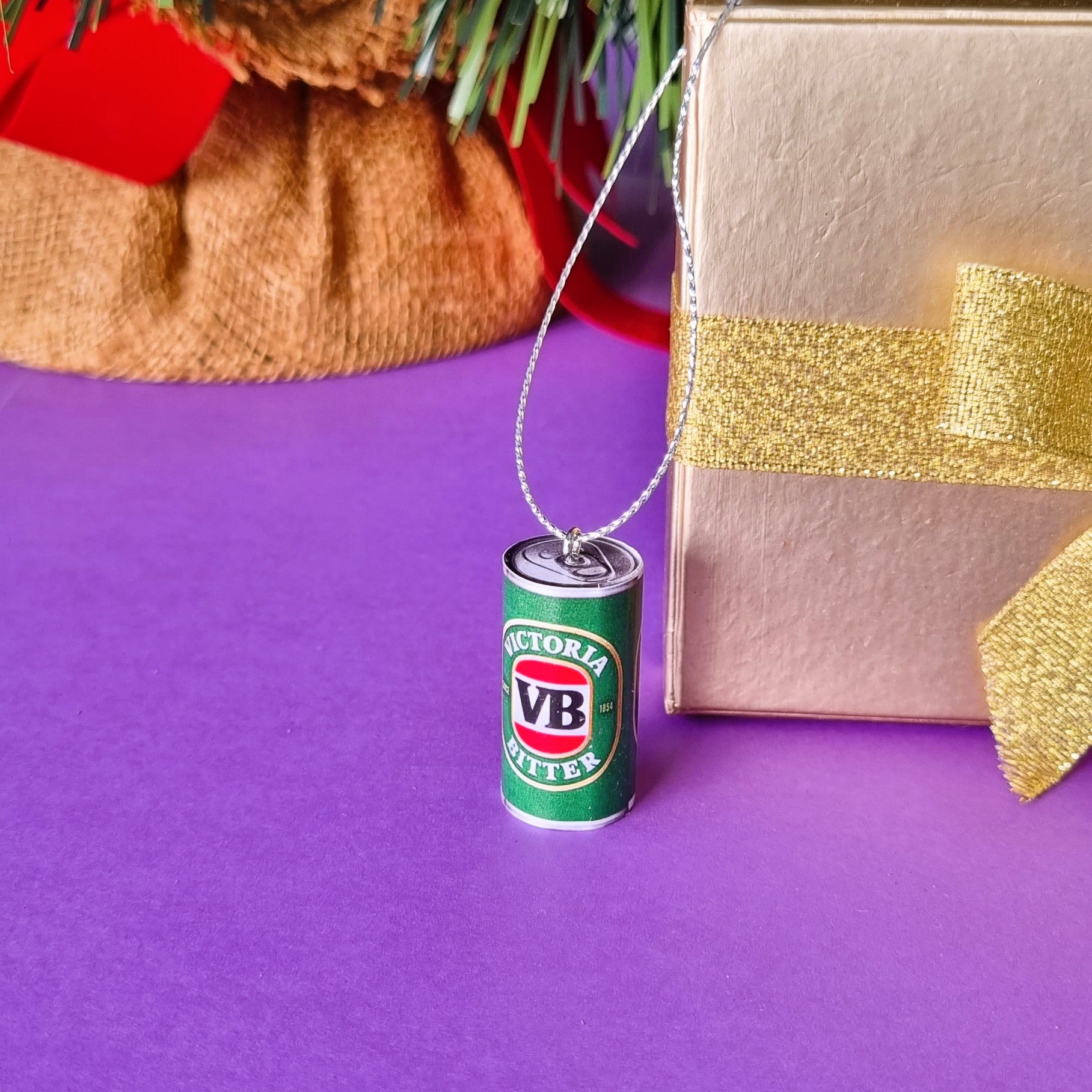 A miniature green and white VB Bitter beer can Christmas ornament standing in front of a golg box prop on a purple surface.