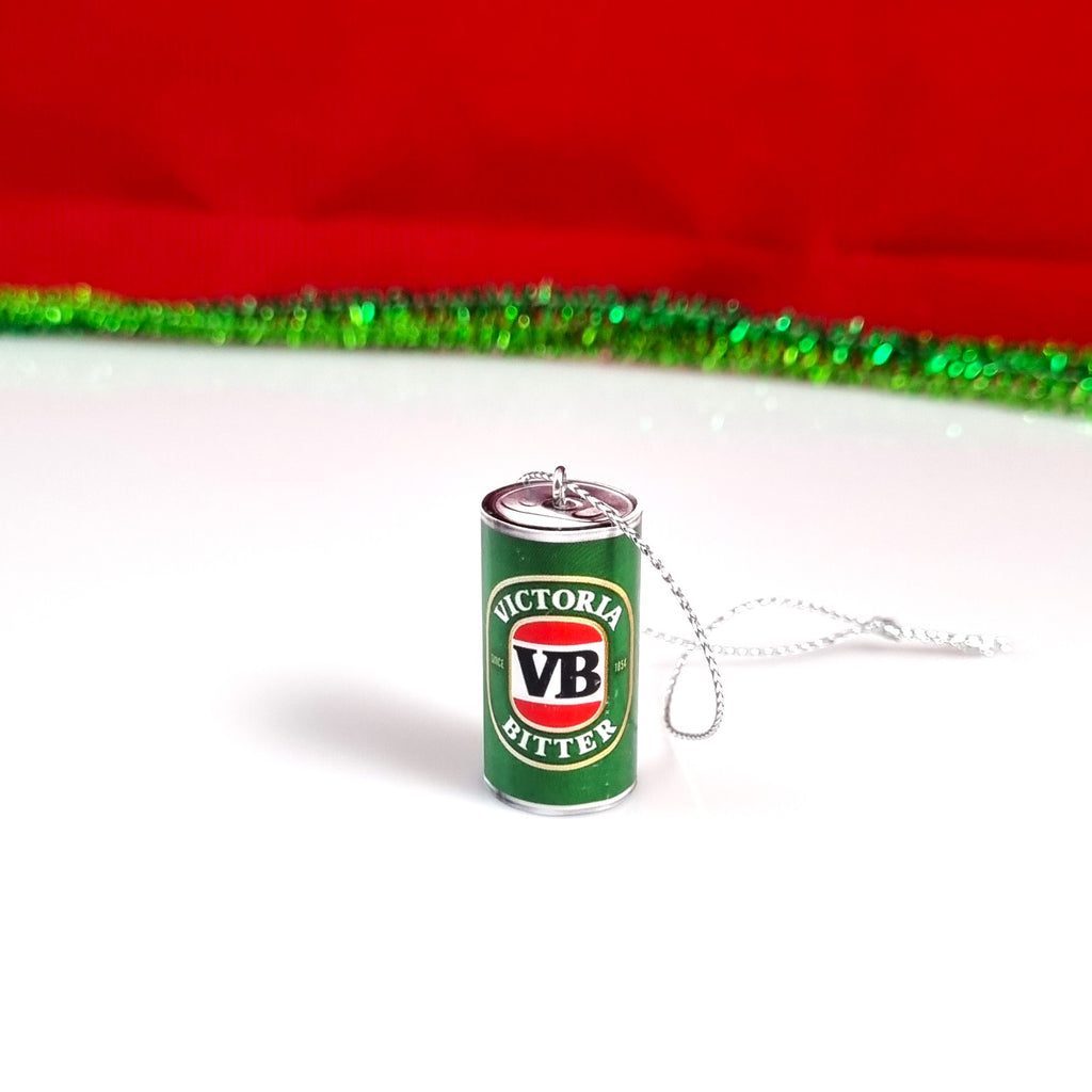 A miniature silver can-shaped Christmas tree ornament featuring the green, red, and white Victoria Bitter logo, resting on a white surface with a red and green tinsel backdrop.