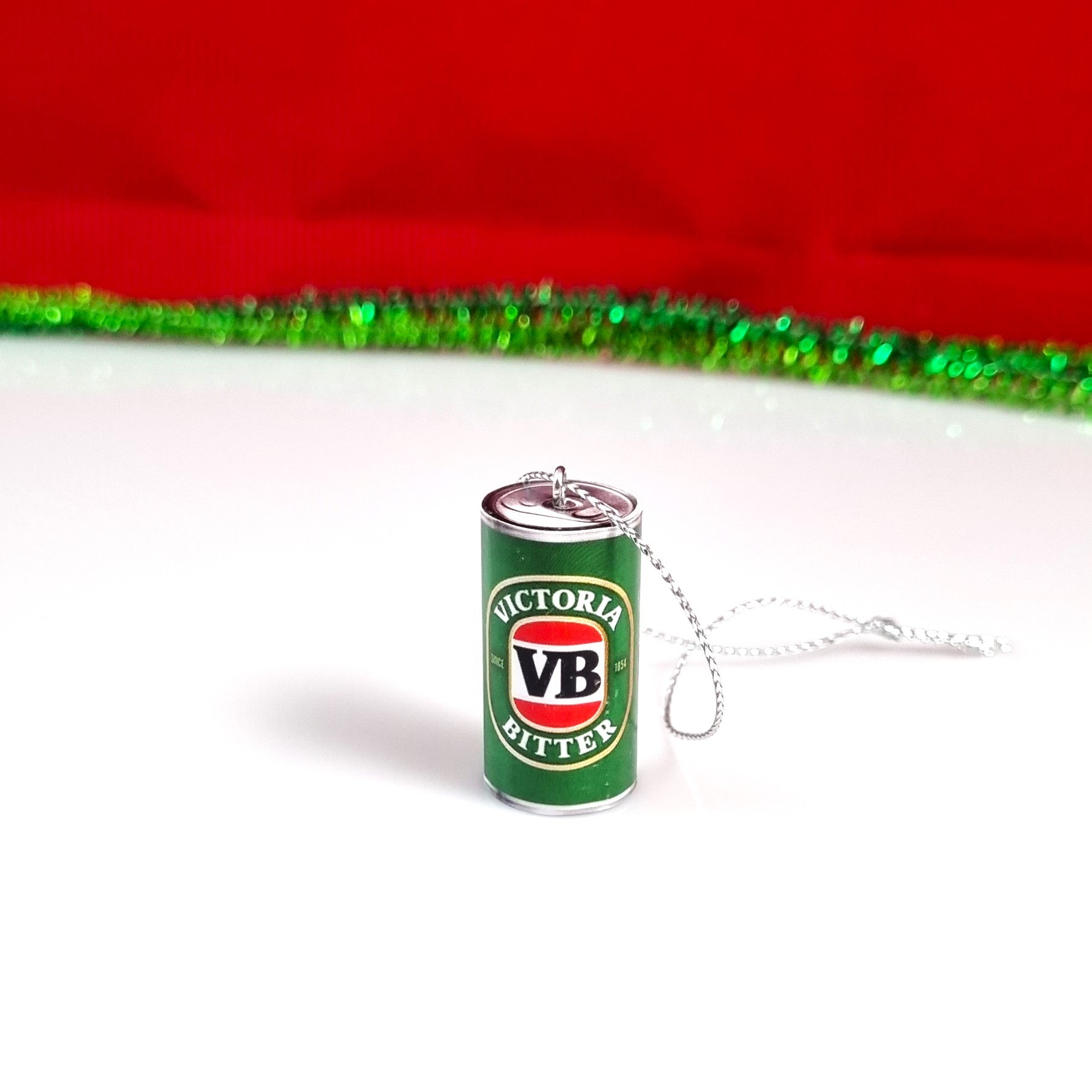 A miniature silver can-shaped Christmas tree ornament featuring the green, red, and white Victoria Bitter logo, resting on a white surface with a red and green tinsel backdrop.