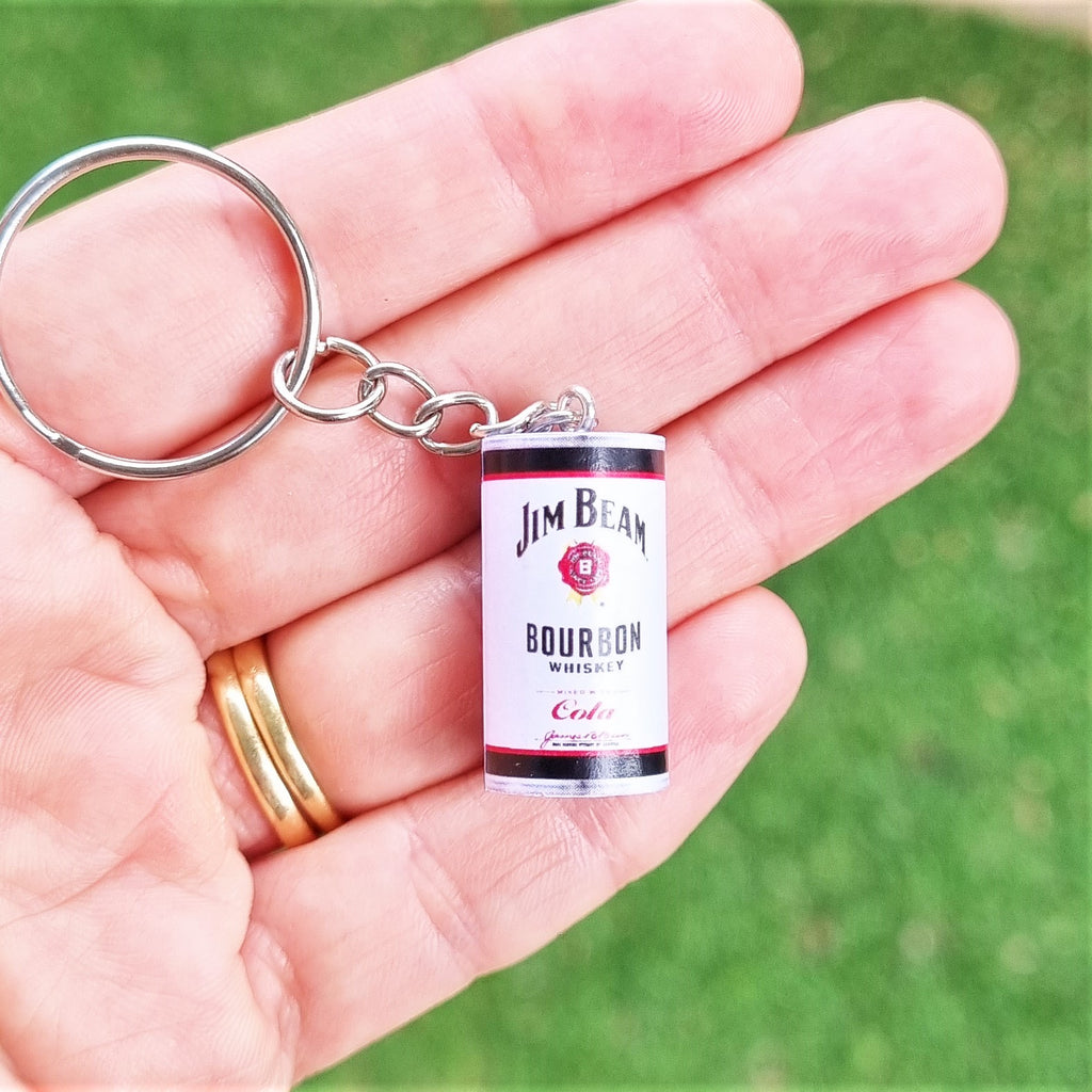 A miniature Jim Beam Bourbon Whiskey keychain held in a person's hand, with a blurred green grass background.