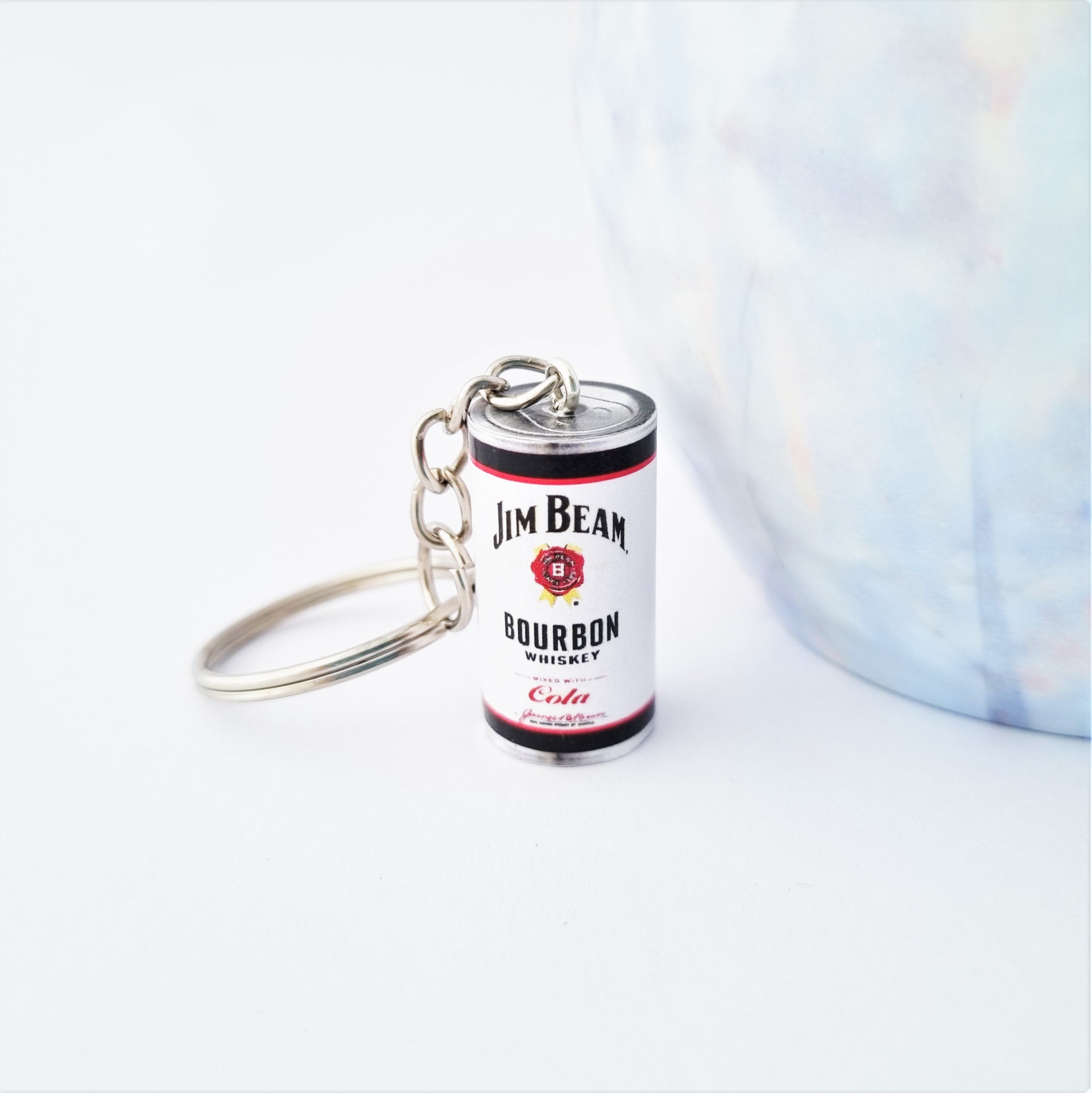 A miniature Jim Bean Bourbon Whiskey keychain shaped like a whiskey can, featuring a silver metal key ring and chain, set against a clean white background with a soft-focus blue and white marble prop on the right.