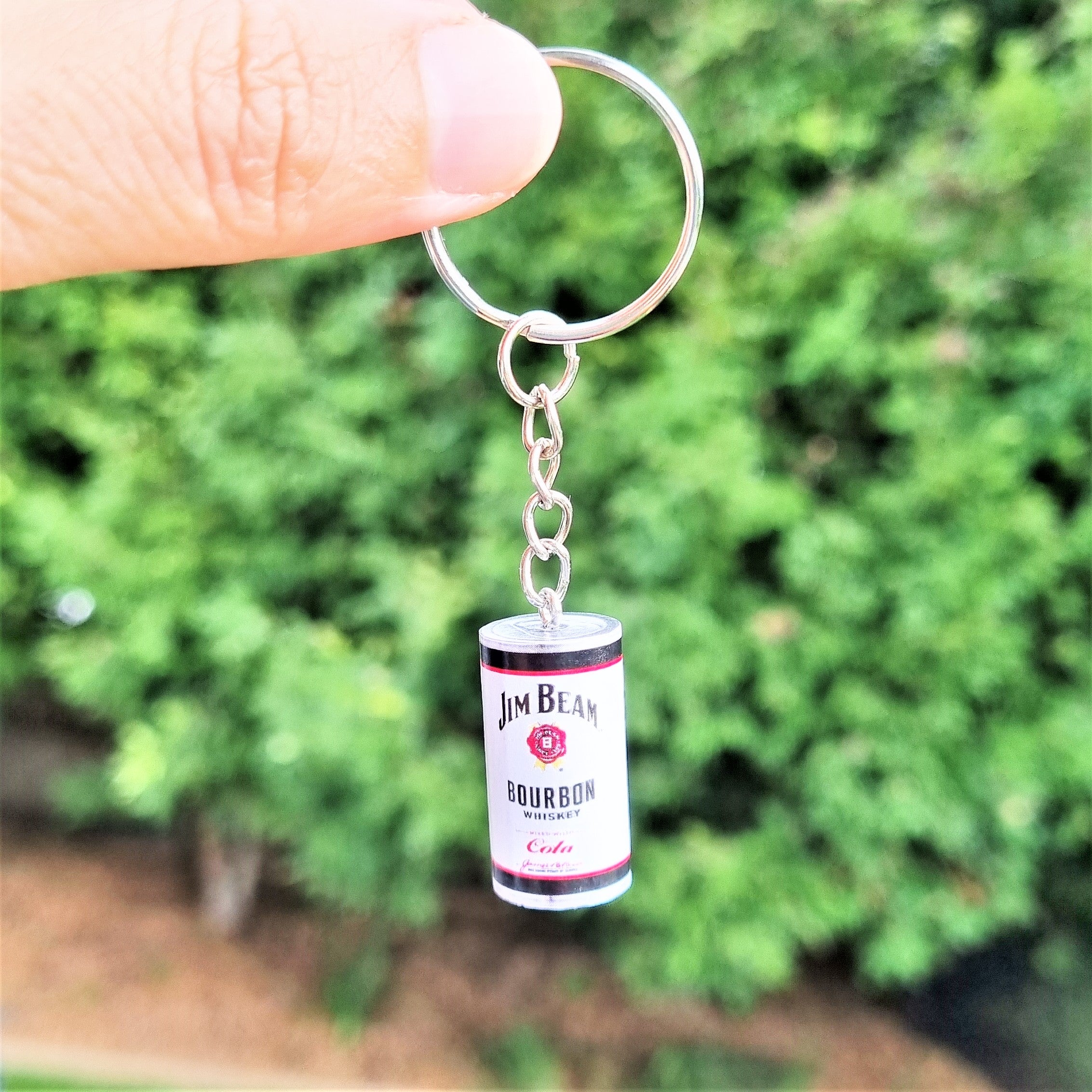A silver metal keychain with a miniature Jim Beam Bourbon and Cola glass can charm, held by a person's finger against a blurred green foliage background.