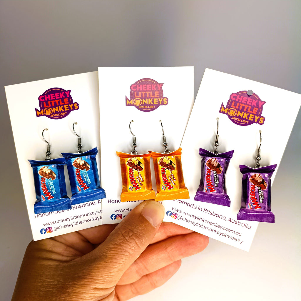 Three pairs of novelty ice cream sandwich earrings displayed on white backing cards, held in a hand. The earrings are designed to look like mini Maxibon ice cream bars in blue, orange, and purple packaging.