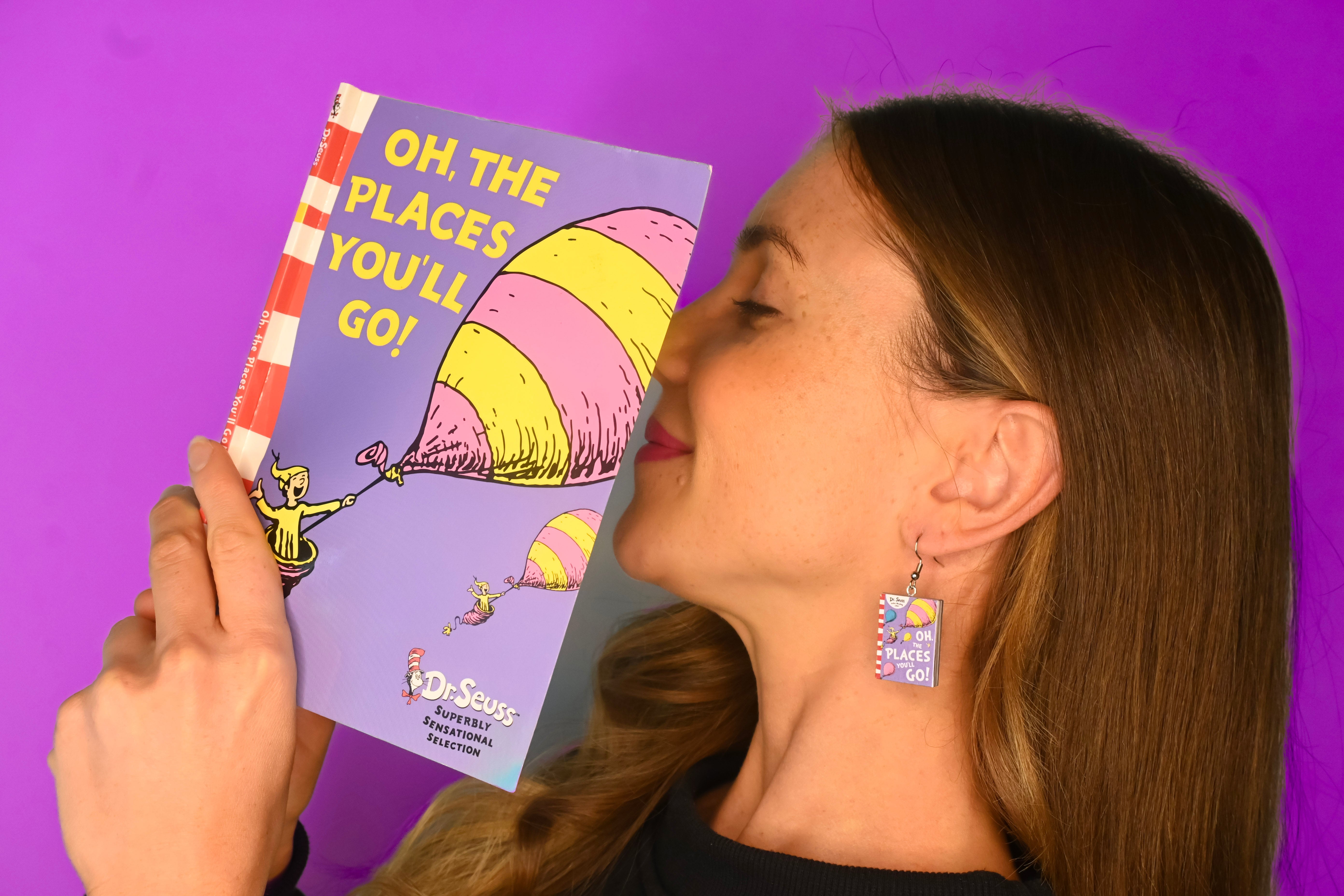 A woman holding a Dr. Seuss 'Oh The Places You'll Go!' book against a solid purple background, wearing miniature Oh The Places You'll Go book earrings.