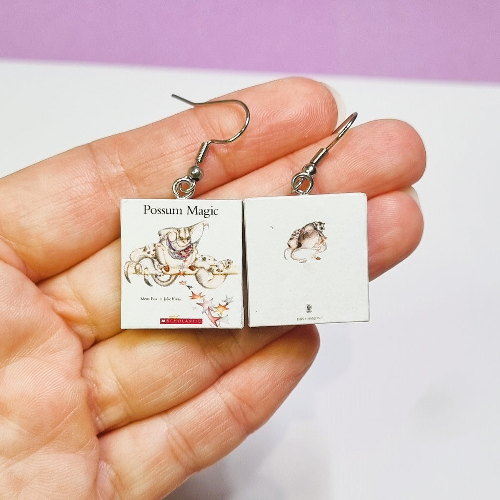 Hand holding a pair of 'Possum Magic' book earrings against a light purple background.