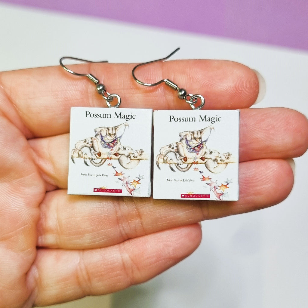 Hand holding earrings with 'Possum Magic' book cover design