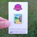 A hand holds a white Cheeky Little Monkeys Jewellery backing card featuring a small book pin of 'The Gruffalo', against an outdoor grassy background.