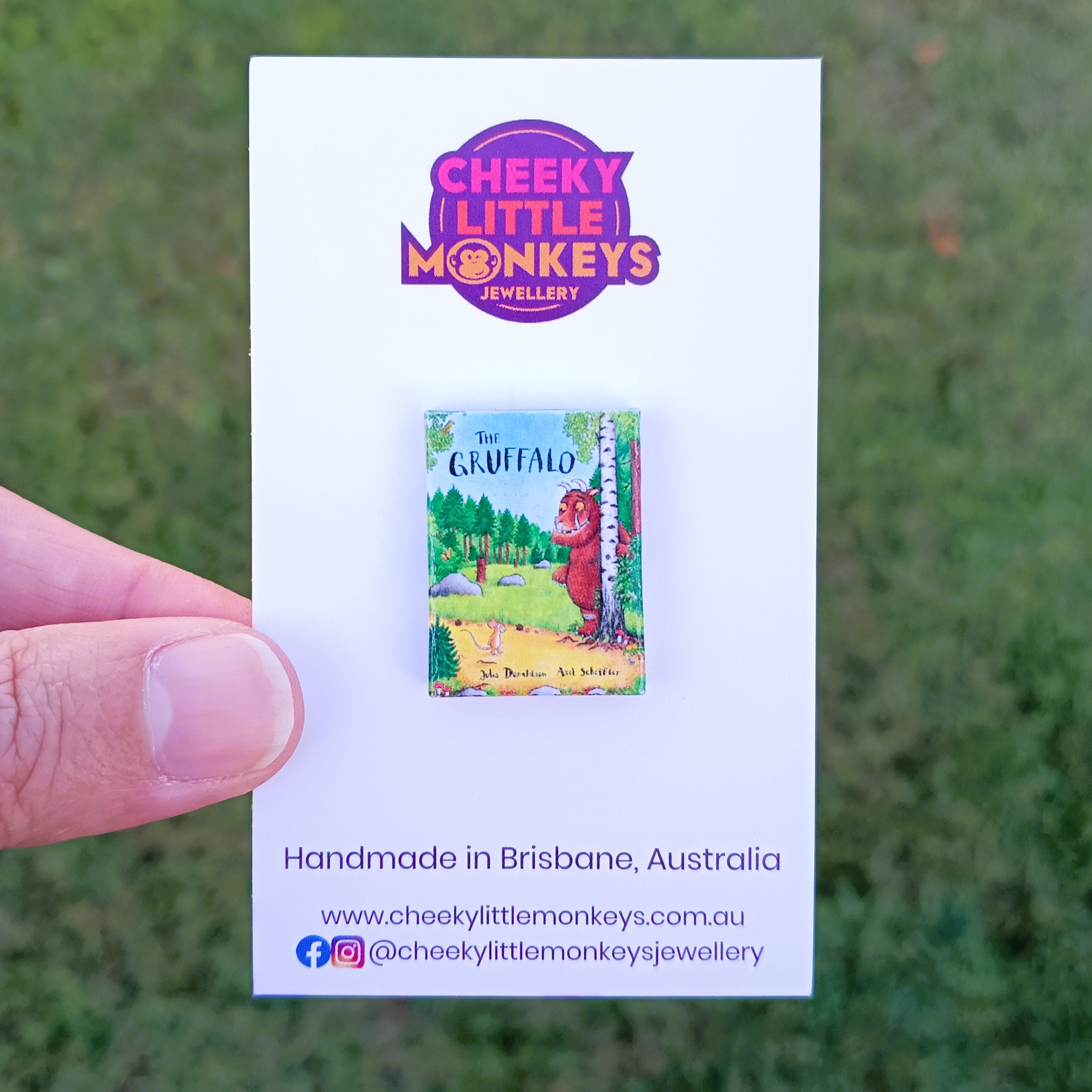 A hand holds a white Cheeky Little Monkeys Jewellery backing card featuring a small book pin of 'The Gruffalo', against an outdoor grassy background.