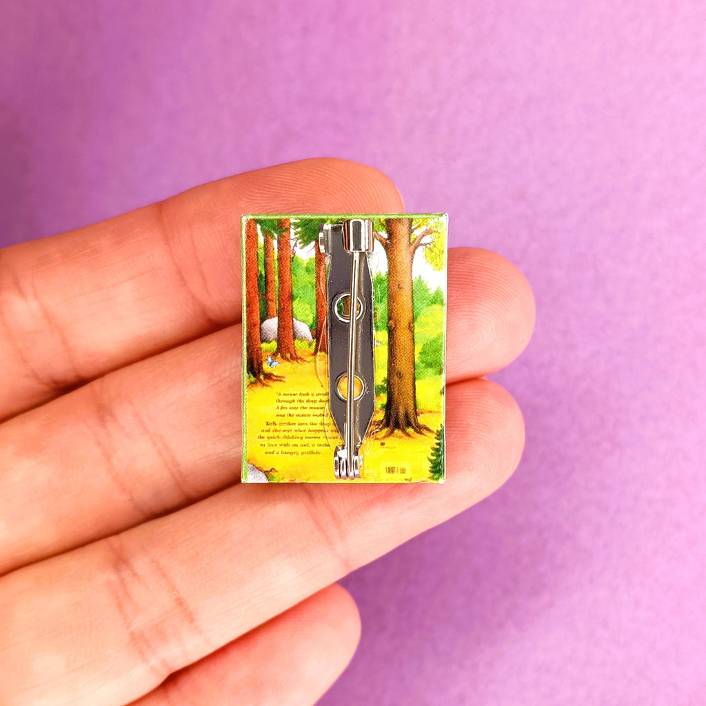 A hand holds a miniature book brooch pin, against a purple background. The book is the back cover of The Gruffalo.