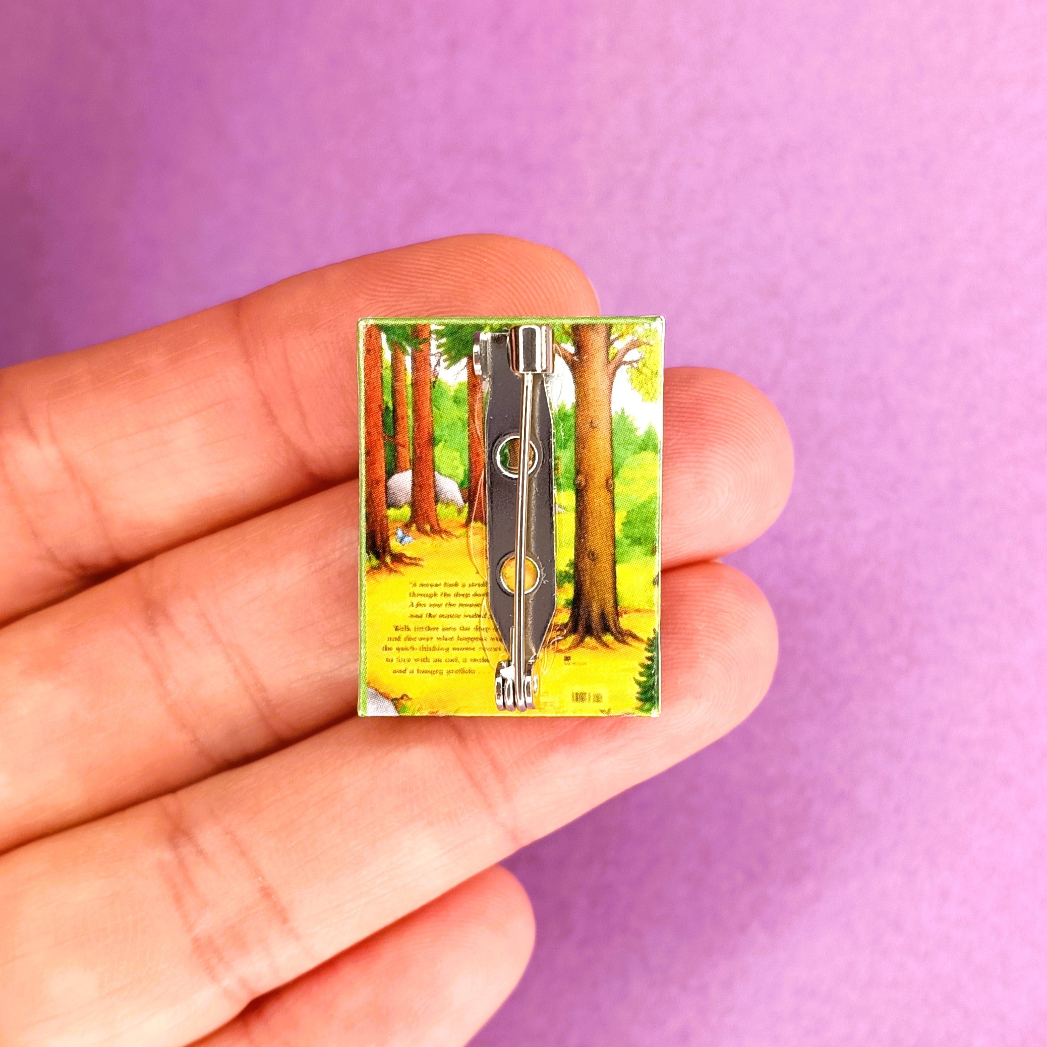 A hand holds a miniature book brooch pin, against a purple background. The book is the back cover of The Gruffalo.