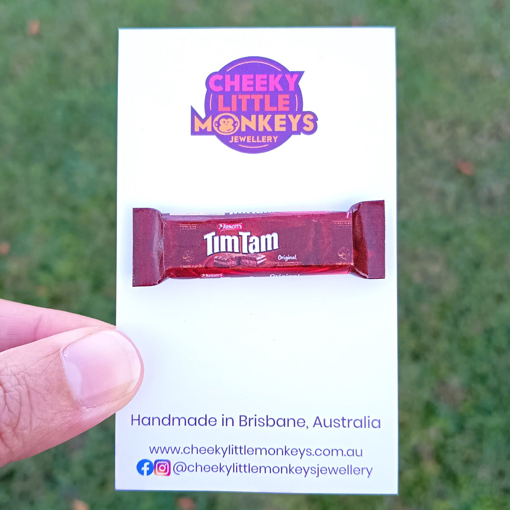 A hand holding a white card featuring a miniature Tim Tam packet replica and the Cheeky Little Monkeys Jewellery logo, with a blurred green outdoor background.