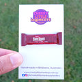 A hand holding a white card featuring a miniature Tim Tam packet replica and the Cheeky Little Monkeys Jewellery logo, with a blurred green outdoor background.