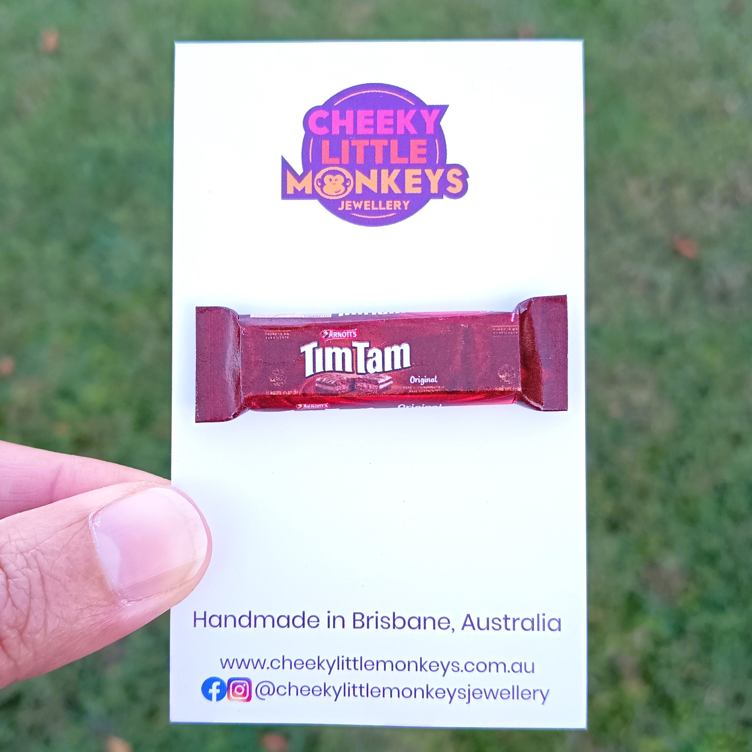 A hand holding a white card featuring a miniature Tim Tam packet replica and the Cheeky Little Monkeys Jewellery logo, with a blurred green outdoor background.