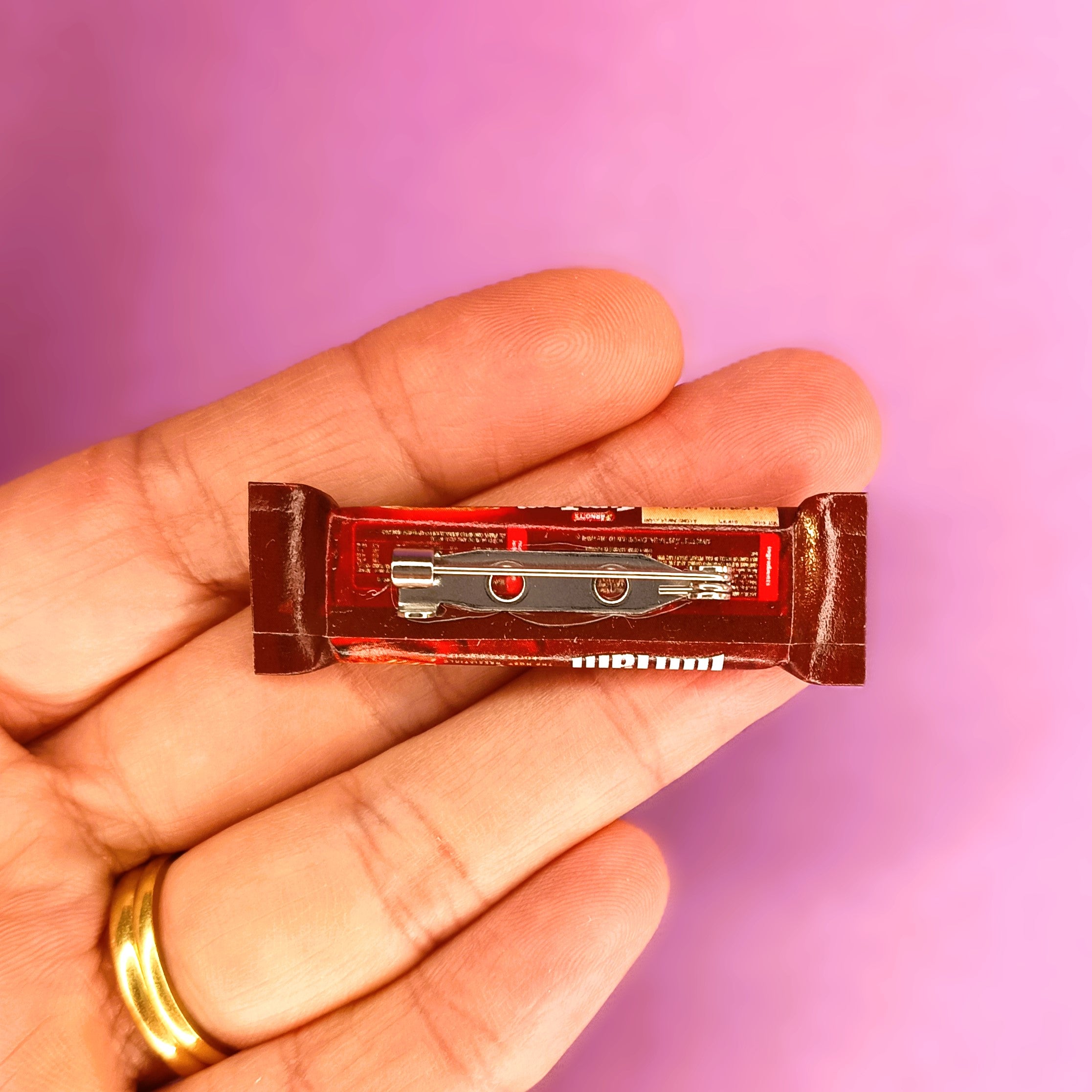 A miniature Tim Tam packet replica with a brooch pin, held in a person's hand against a solid pink background.