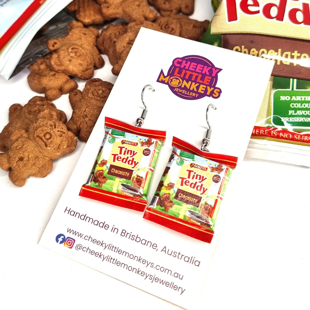 A pair of dangle earrings shaped like miniature 'Tiny Teddy' chocolate snack bags, displayed on a branded white backing card. The card is styled on a white surface with chocolate teddy bear cookies and a bag of 'Tiny Teddy' chocolates in the background.