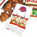 A pair of dangle earrings shaped like miniature 'Tiny Teddy' chocolate snack bags, displayed on a branded white backing card. The card is styled on a white surface with chocolate teddy bear cookies and a bag of 'Tiny Teddy' chocolates in the background.