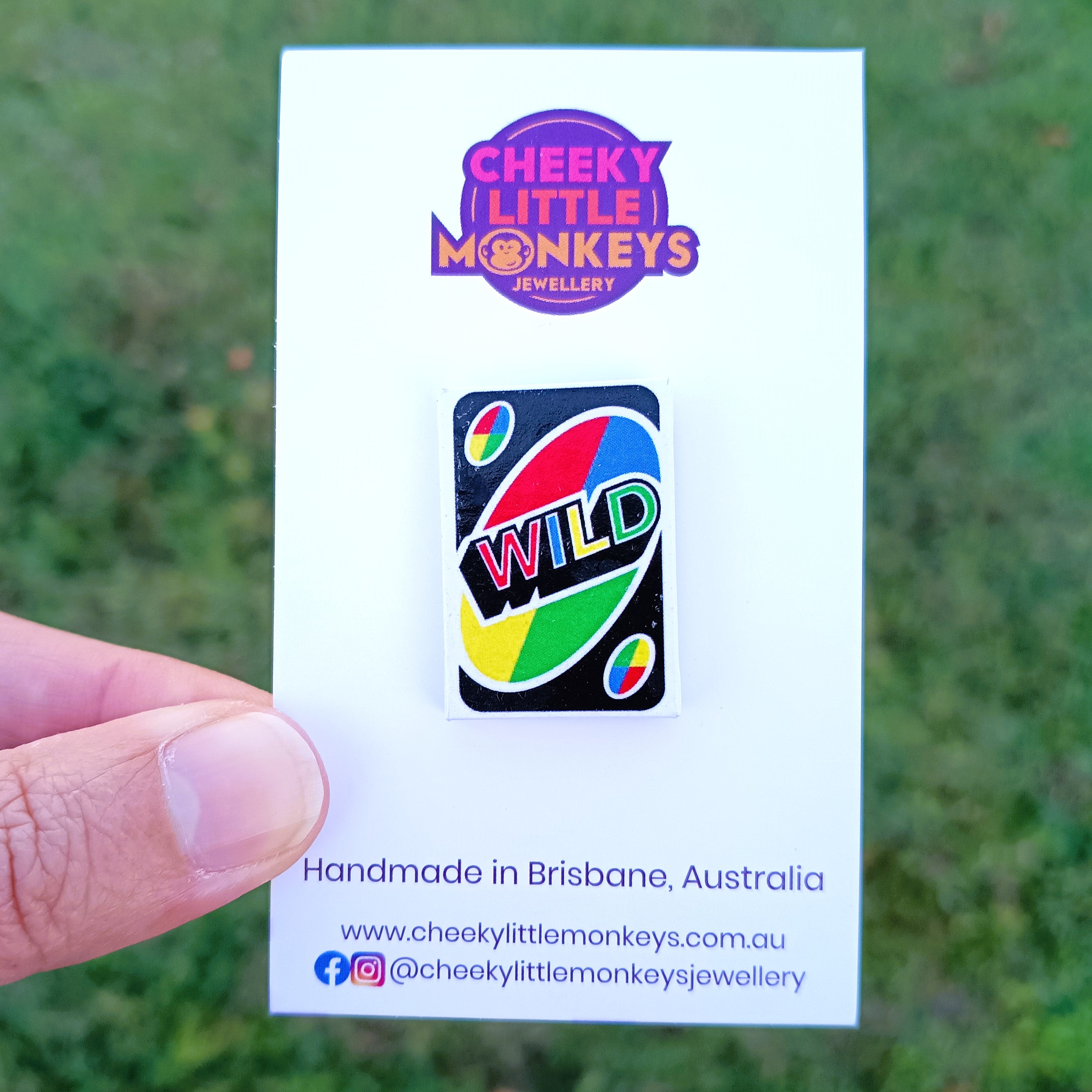 A hand holds a white backing card featuring a 'Cheeky Little Monkeys Jewellery' pin. The pin is shaped like a UNO 'WILD' card with a black background and red, yellow, green, and blue curved graphics.