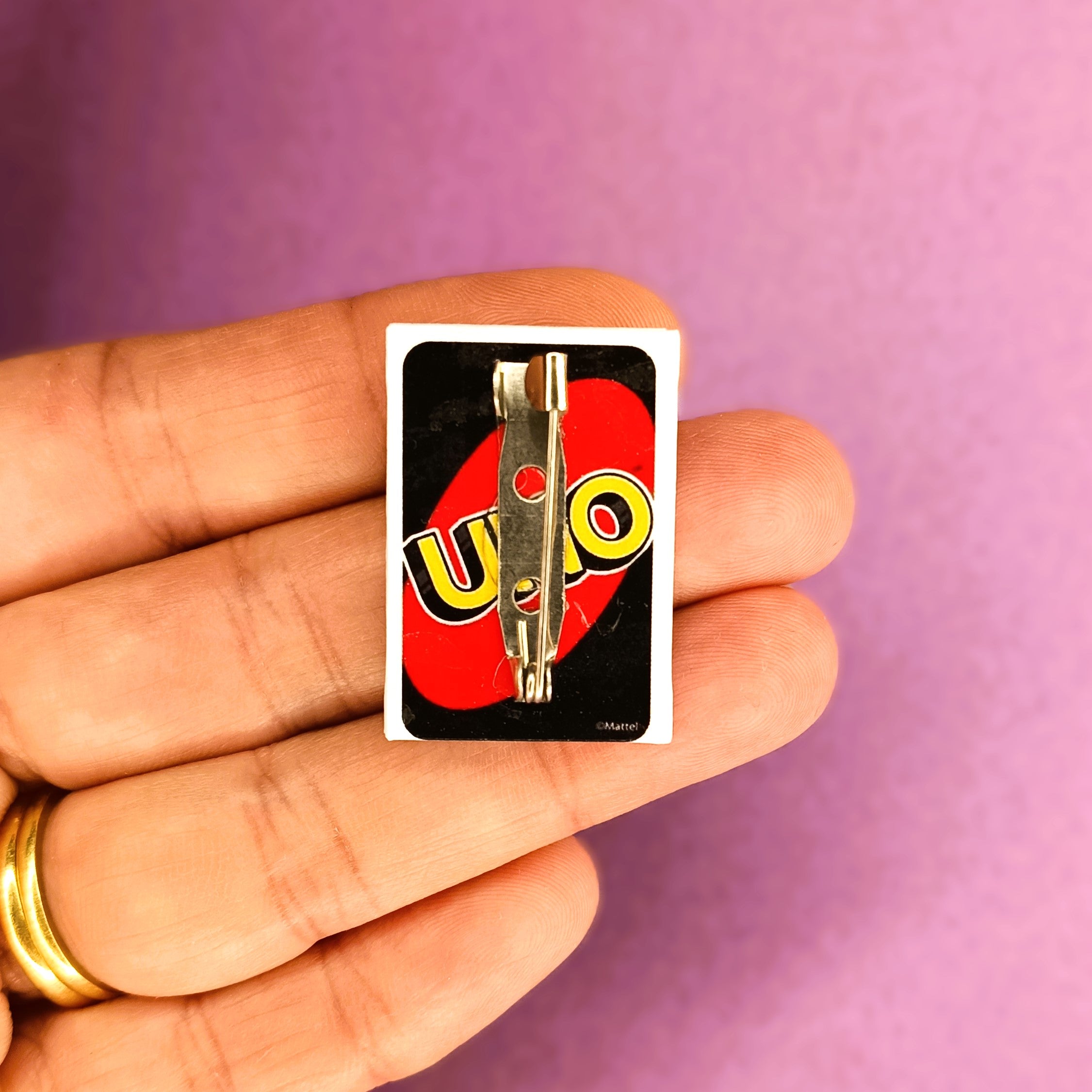 A small UNO card pin or badge is held on a person's fingers against a solid purple background. The badge features the classic black, red and yellow UNO card.