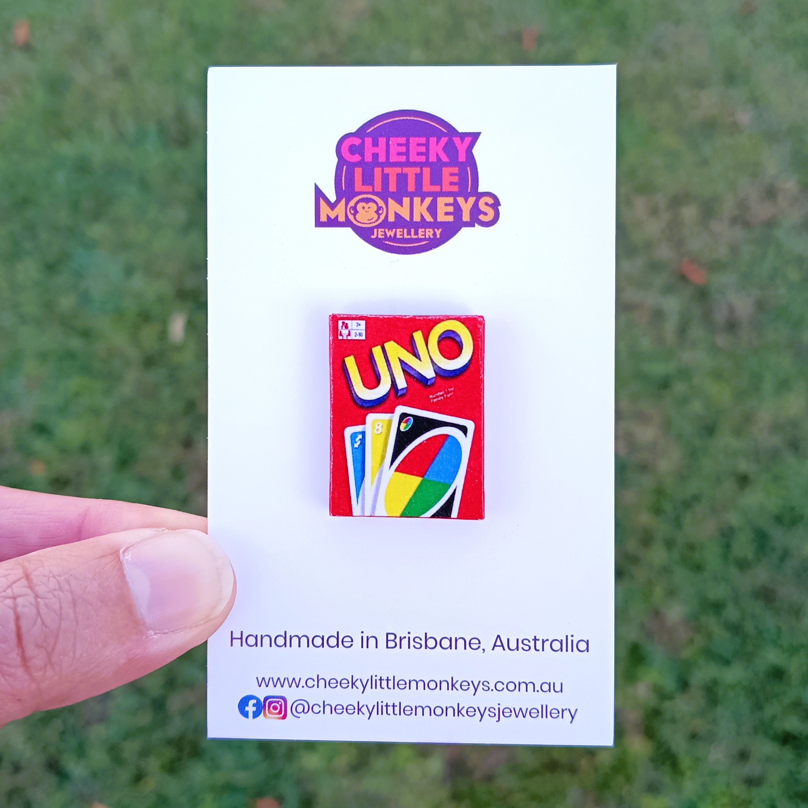 A hand holds a white backing card featuring a small UNO-themed pin. The card shows the brand logo 'Cheeky Little Monkeys Jewellery' and text reading 'Handmade in Brisbane, Australia' against a blurred green grass background.