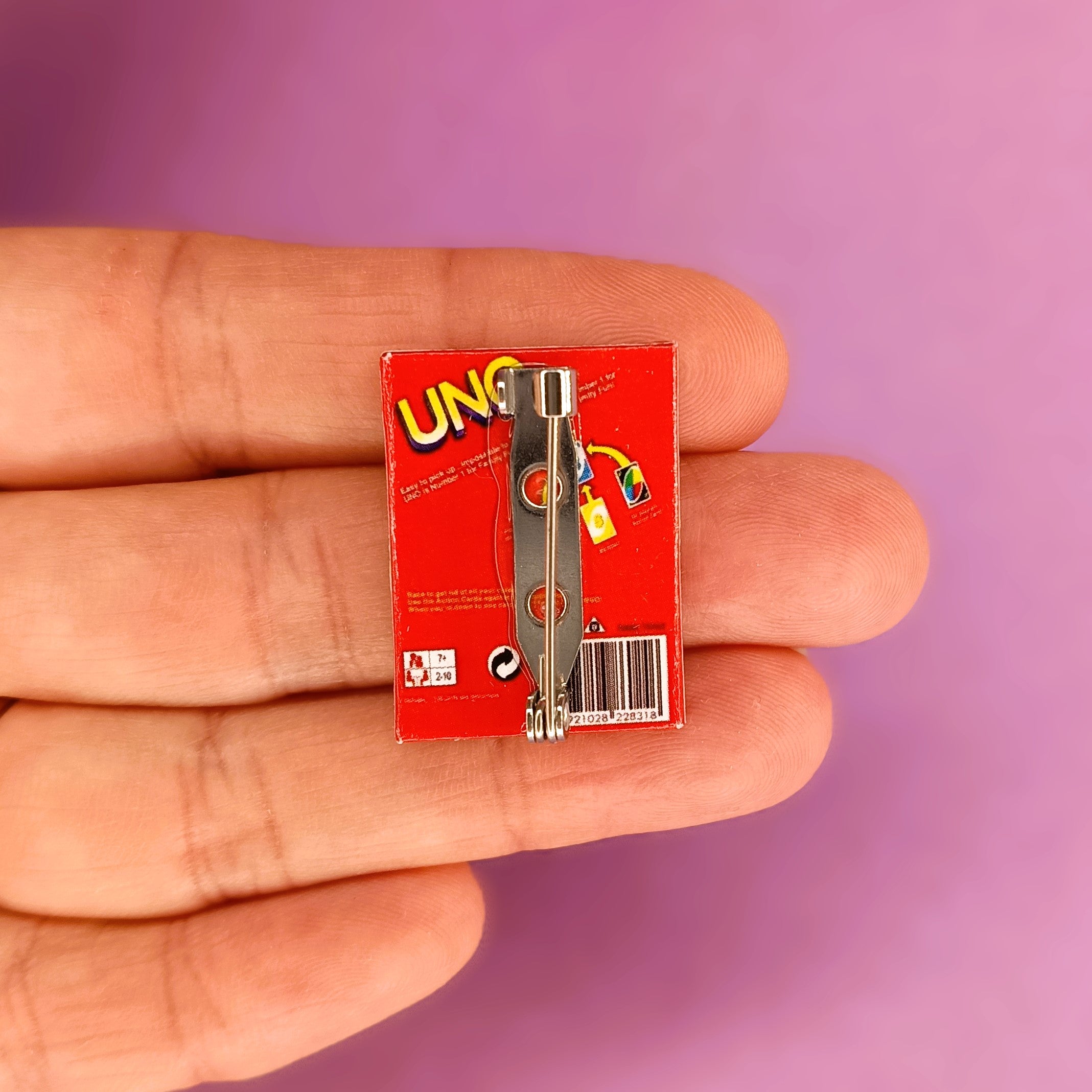 A miniature metal UNO pin or brooch on its back, held in a person's hand against a solid purple background