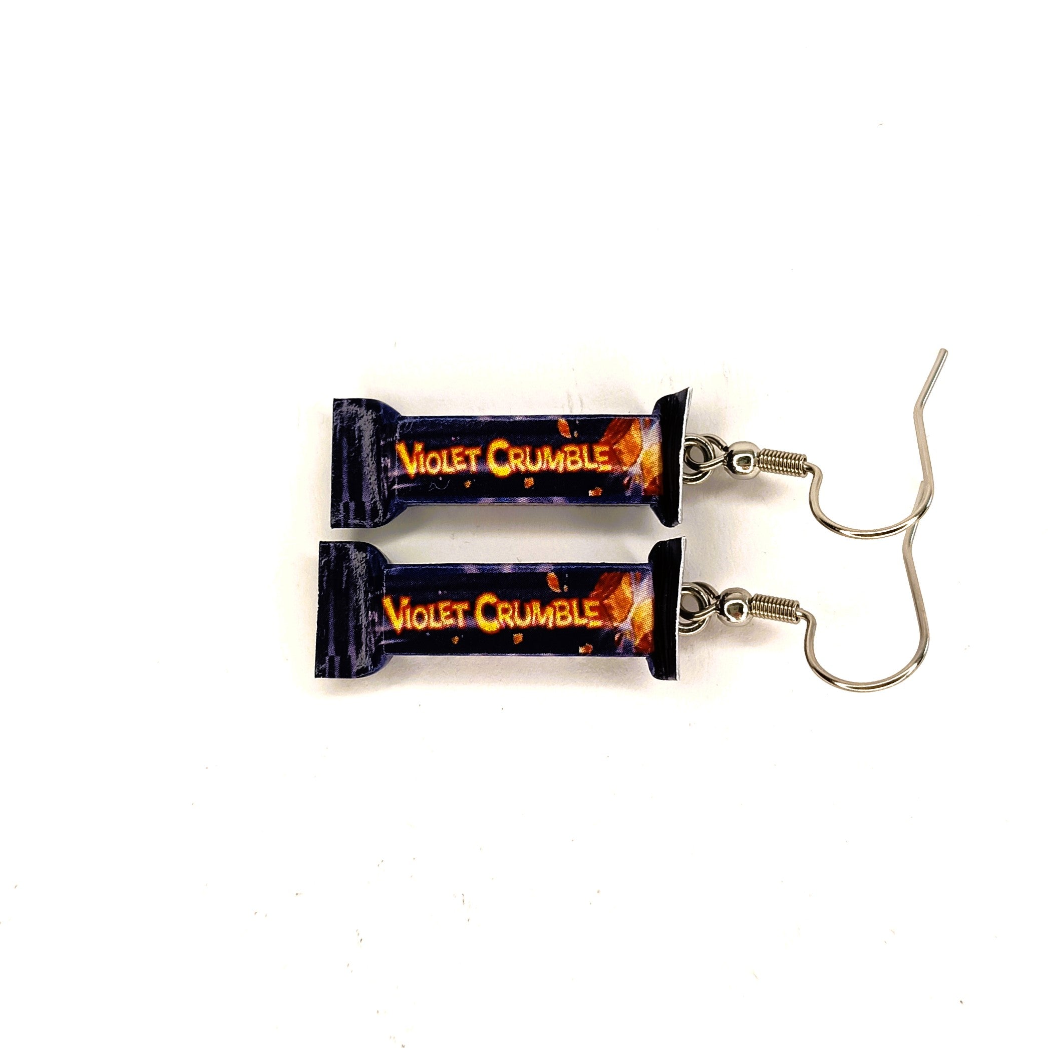 A pair of dangling earrings shaped like wrapped candy bars, printed with the text 'Violet Crumble' in orange on a dark purple background with small orange details. The earrings have silver fishhook wires and are displayed on a plain white background.