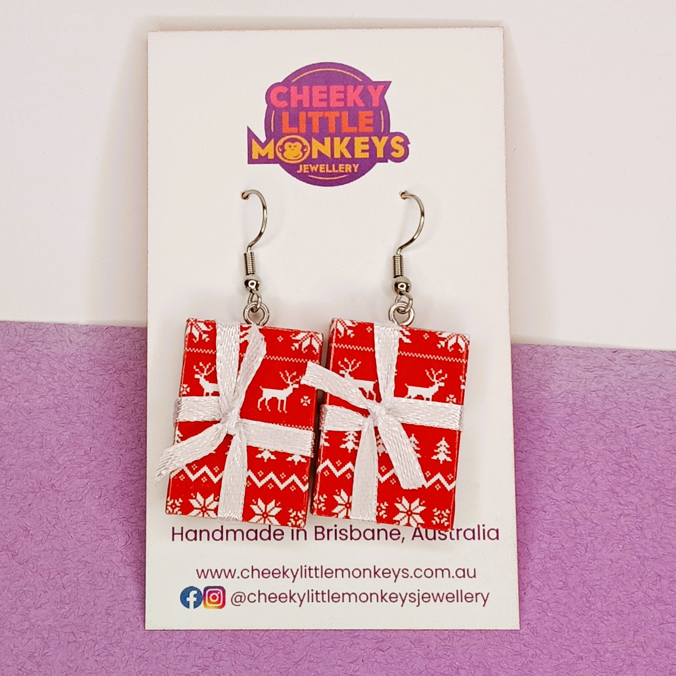 A pair of rectangular dangle earrings shaped like wrapped Christmas presents, displayed on a white branded card. The earrings have red wrapping paper with white snowflake and deer patterns, tied with white ribbon, and hang from silver hooks.