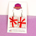 A pair of handmade rectangular dangle earrings by Cheeky Little Monkeys Jewellery, featuring a red and white Christmas tree pattern wrapped in a red ribbon bow, displayed on a branded white backing card against a split white and purple background.