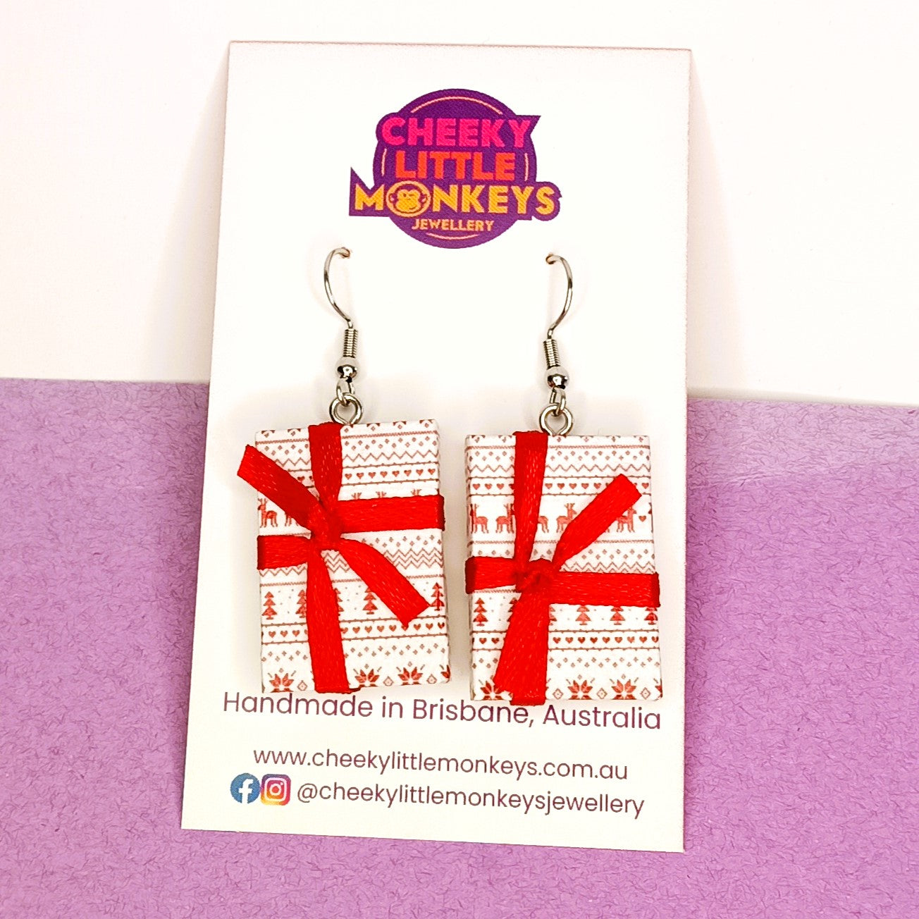A pair of handmade rectangular dangle earrings by Cheeky Little Monkeys Jewellery, featuring a red and white Christmas tree pattern wrapped in a red ribbon bow, displayed on a branded white backing card against a split white and purple background.