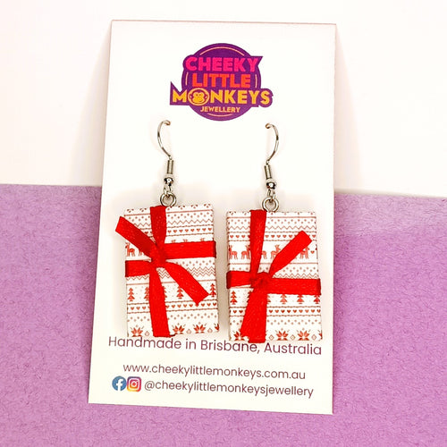 A pair of handmade rectangular dangle earrings by Cheeky Little Monkeys Jewellery, featuring a red and white Christmas tree pattern wrapped in a red ribbon bow, displayed on a branded white backing card against a split white and purple background.